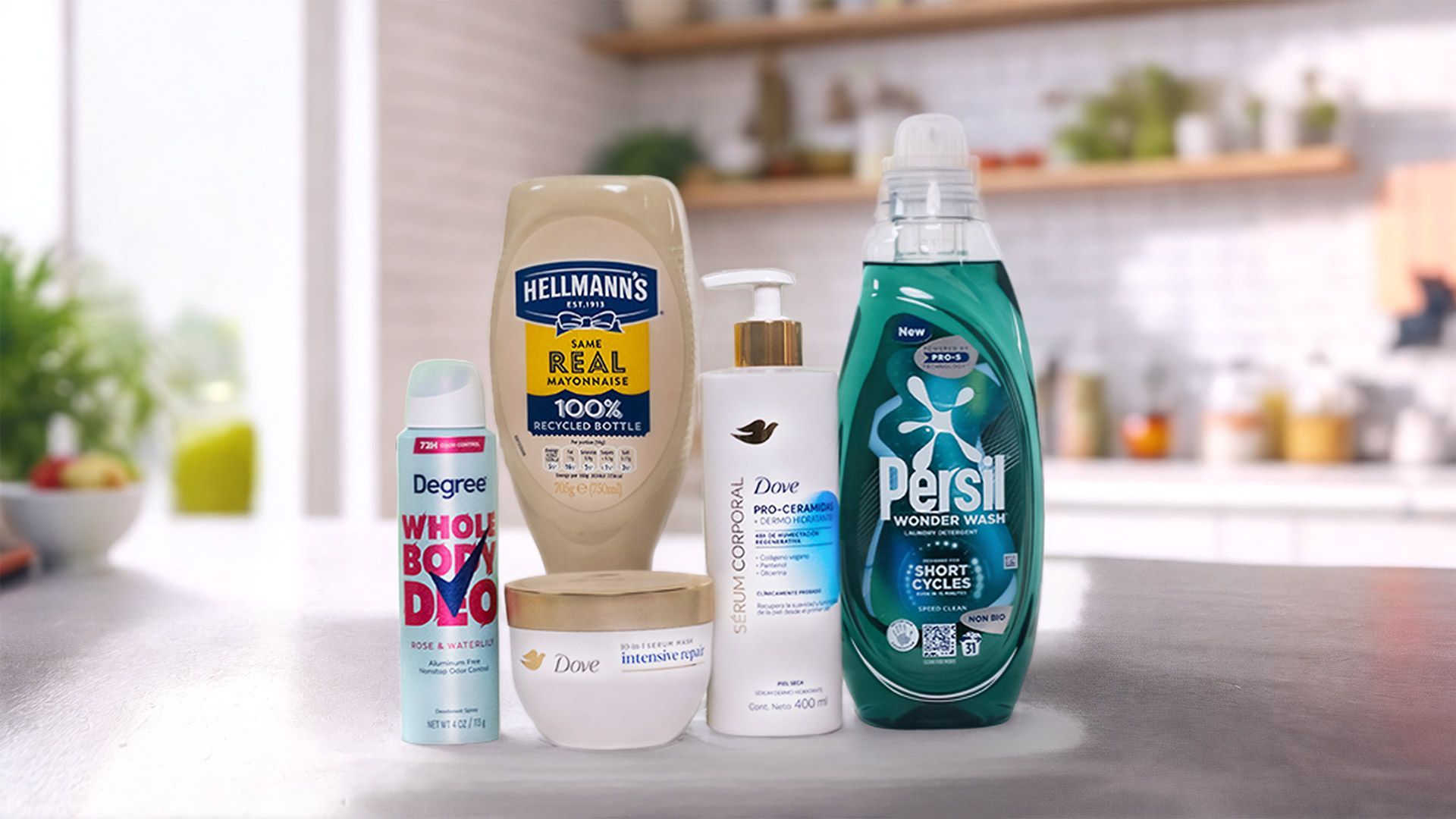 A variety of Unilever's Power Brands arranged on a kitchen counter. From left to right, you can see a jar of Knorr chicken bouillon, a Degree deodorant spray labelled "WHOLE BODY DEO", a box of Magnum ice cream labelled "UTOPIA", a bottle of Dove body lotion pro-ceramides serum, a jar of Dove Intensive Repair cream and a bottle of Persil Wonder Wash. In the background, there's a modern kitchen with plants and various items on the shelves.
